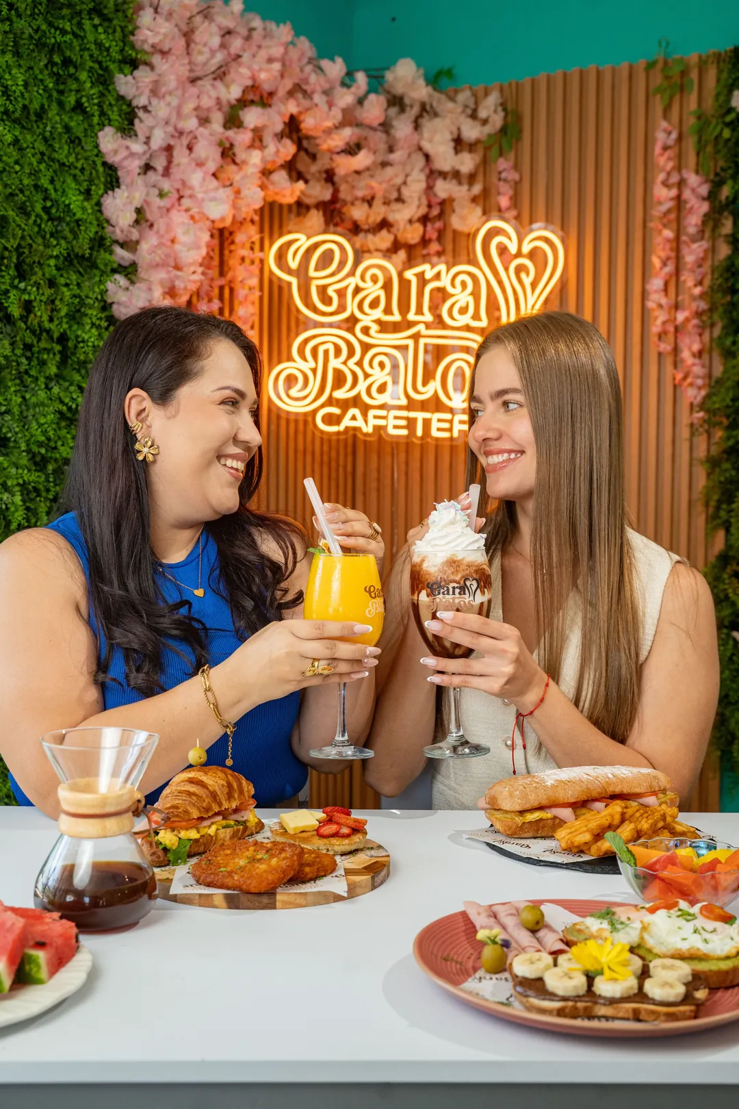 Two friends toast with drinks in front of the Garabatos neon sign